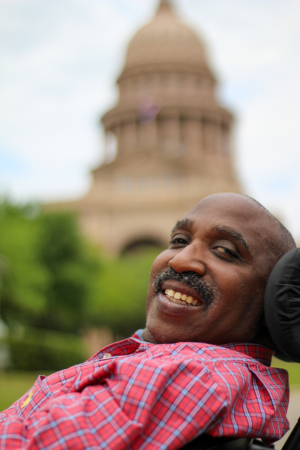 An image of Ricky Broussard, who is smiling and seated in his power wheelchair. He is wearing a red plaid shirt with a collar, and the Texas Capitol dome is in the distant background.